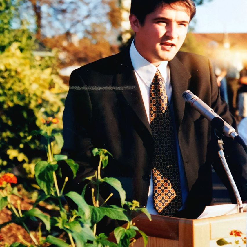 Ein Mann in formeller Kleidung steht an einem Rednerpult und spricht in ein Mikrofon, mit einem Garten im Hintergrund.