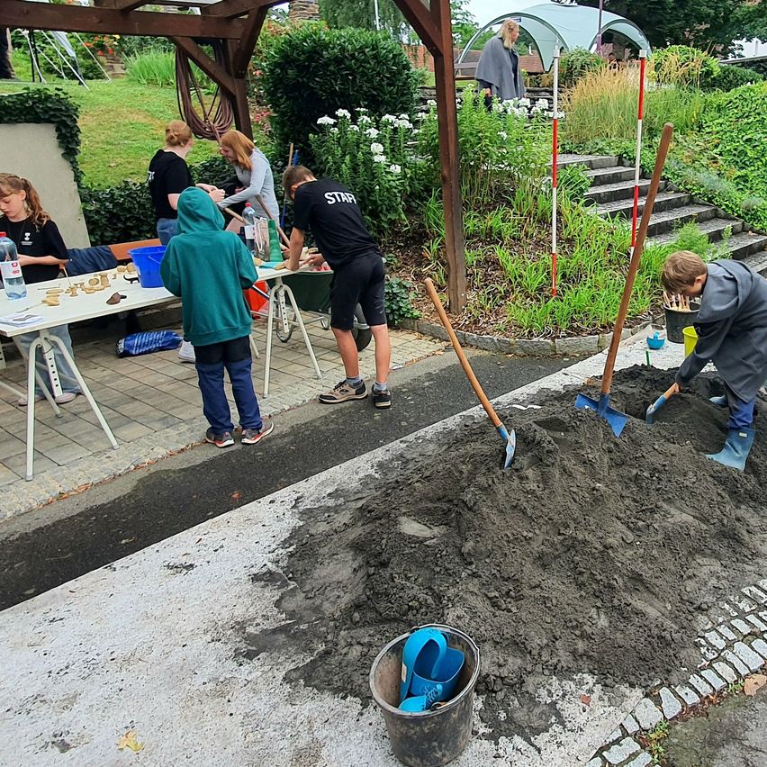 Mehrere Personen arbeiten in einem Gartenbereich. Ein Junge gräbt, während ein weiteres Kind und zwei Frauen an einem Tisch sitzen. In der Nähe steht eine Frau in der Nähe eines Zeltes und einer Treppe.