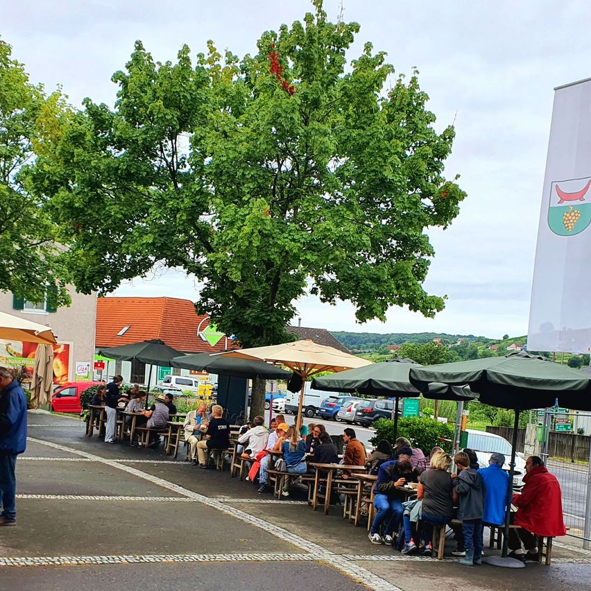 Ein Freiluft-Essbereich mit Tischen und Stühlen unter Sonnenschirmen, Menschen sitzen und unterhalten sich. Der Bereich ist von Bäumen und Gebäuden umgeben. In der Ferne sind Autos geparkt.