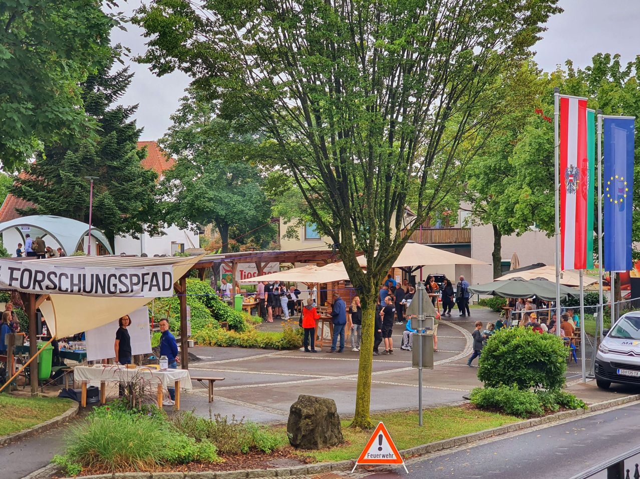 Freiluftmarkt mit Ständen und Sonnenschirmen. Menschen gehen herum, einige sitzen an Tischen. Ein Baum mit einem Warnschild für Feuergefahr ist sichtbar.
