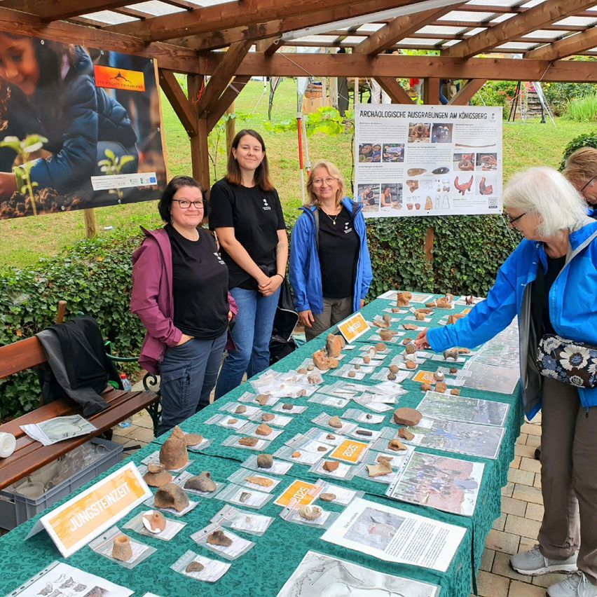Eine Gruppe von Frauen steht unter einem hölzernen Pavillon, möglicherweise an einem archäologischen Ausgrabungsort. Vor ihnen ist ein Tisch mit verschiedenen Steinen und Papieren aufgebaut. Die Frau auf der rechten Seite zeigt auf die Steine. Hinter ihnen ist ein Plakat am Pavillon befestigt.