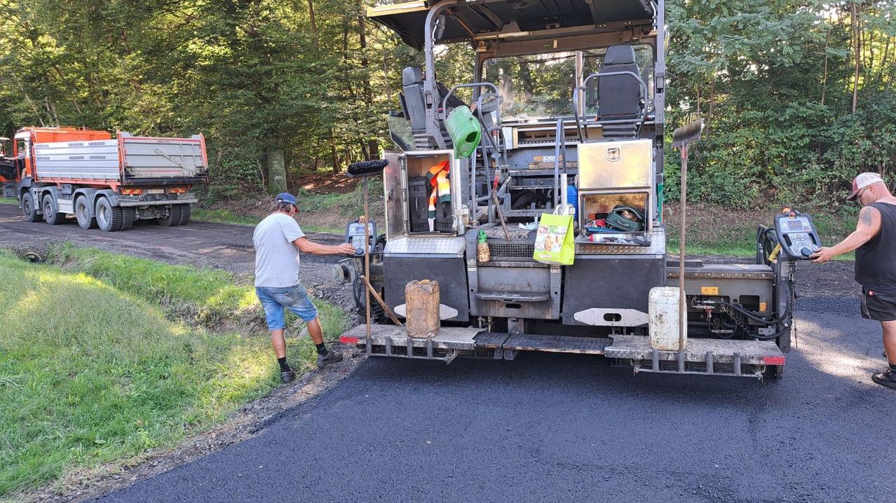 Ein Mann benutzt ein Werkzeug, um den Asphalt auf der Straße zu glätten. Dahinter steht eine Maschine mit eingelagerten Gegenständen. Im Hintergrund befinden sich Bäume und Gras.