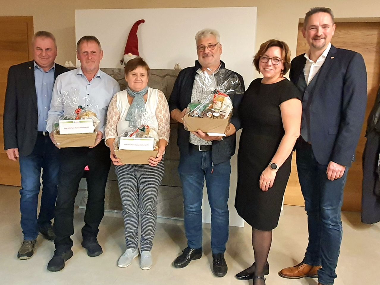 Fünf Menschen stehen in einer Reihe, lächeln und posieren für ein Foto. Sie halten Geschenkboxen mit verschiedenen Gegenständen. Der Hintergrund zeigt eine weiße Wand mit einem Weihnachtszwerg.