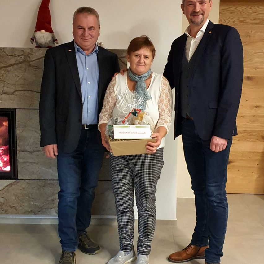 Drei Personen stehen drinnen; eine Frau hält eine Geschenkbox und zwei Männer stehen neben ihr. Hinter ihnen ist ein Kamin mit einer Gnom-Statue darauf. Die Frau lächelt und die Männer tragen formelle Kleidung.