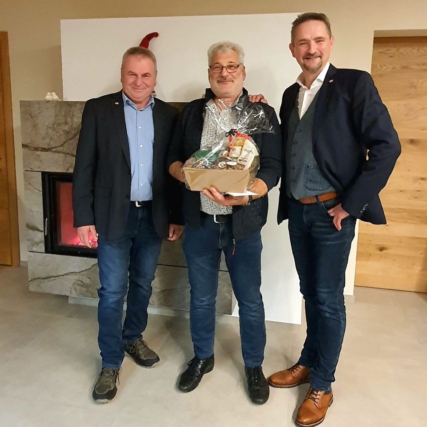 Drei Männer stehen nebeneinander und lächeln, einer hält eine Geschenkbox. Sie stehen vor einem Kamin mit einem Marmorsims.