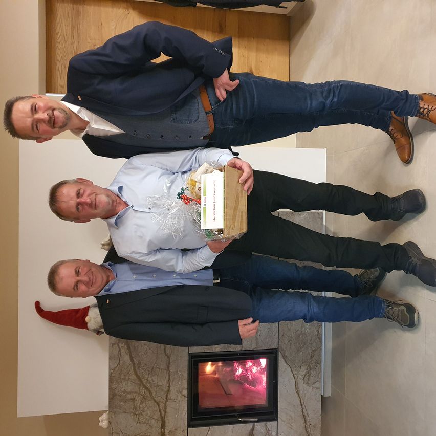 Drei Männer stehen zusammen in einem Raum mit einem Kamin. Der Mann in der Mitte hält eine Box. Die Männer lächeln alle.