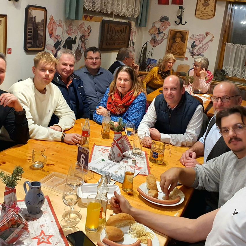Eine Gruppe von Menschen, wahrscheinlich Kollegen, sitzt um einen Tisch in einem Restaurant herum. Sie haben Essen und Getränke vor sich. Der Tisch hat Flaschen, Gläser und Teller. Hinter ihnen hängen Bilderrahmen an der Wand.