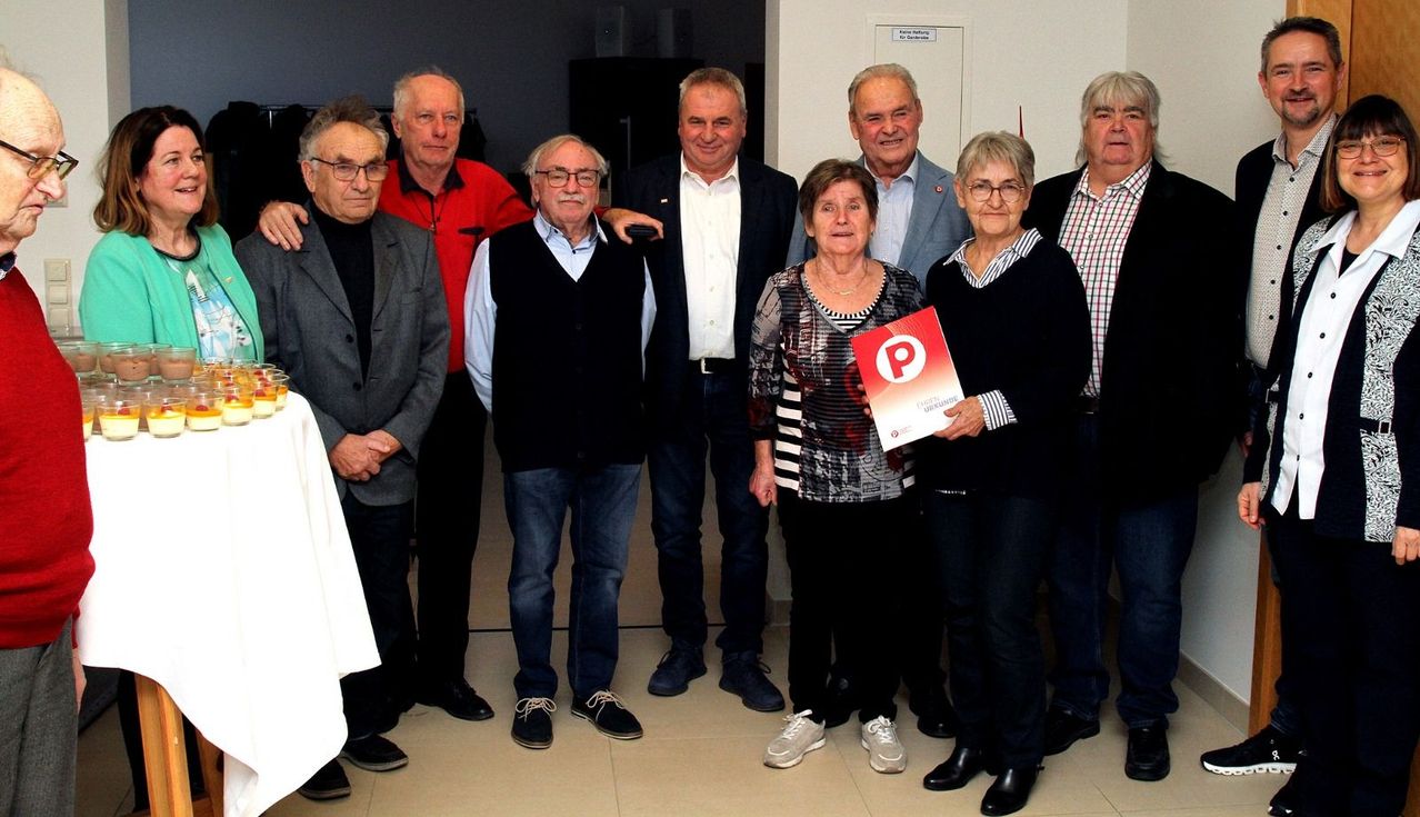 Eine Gruppe älterer Erwachsener, sowohl Männer als auch Frauen, posiert für ein Foto, wobei eine Frau ein rotes Schild hält.