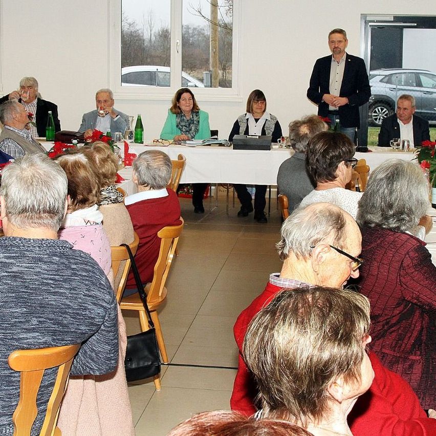 Eine Versammlung älterer Personen, die um einen langen Tisch herum sitzen. Ein Mann steht vorne, möglicherweise spricht er sie an. Flaschen und Gläser sind auf dem Tisch. Hinter ihnen zeigt ein Fenster geparkte Autos.