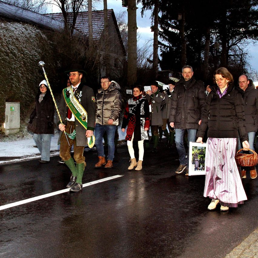 Eine Gruppe von Menschen geht im Winter auf einer nassen Straße, einige in traditionellen Kostümen, andere in Winterkleidung. Sie scheinen an einer Parade teilzunehmen.