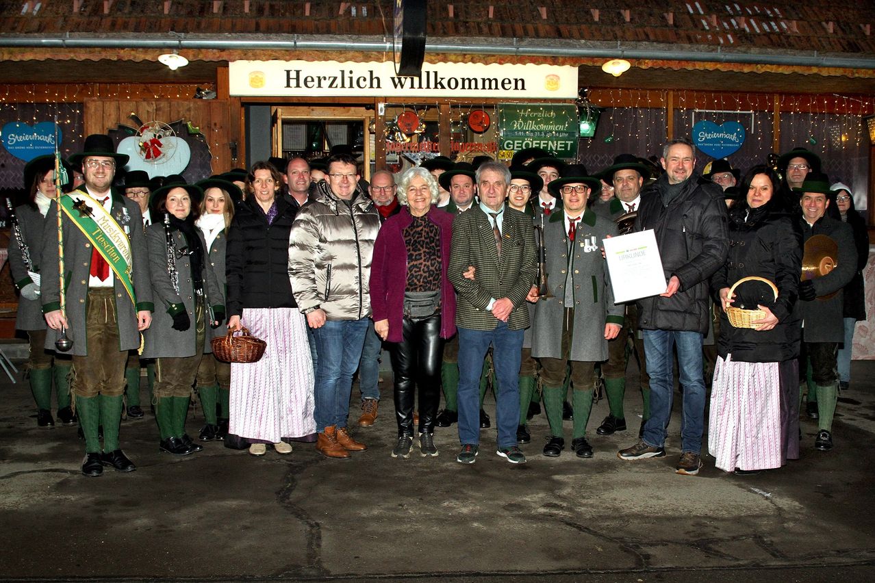 Eine Gruppe von Menschen steht vor einem Restaurant, alle in Winterkleidung. Ein Mann hält ein Zertifikat.