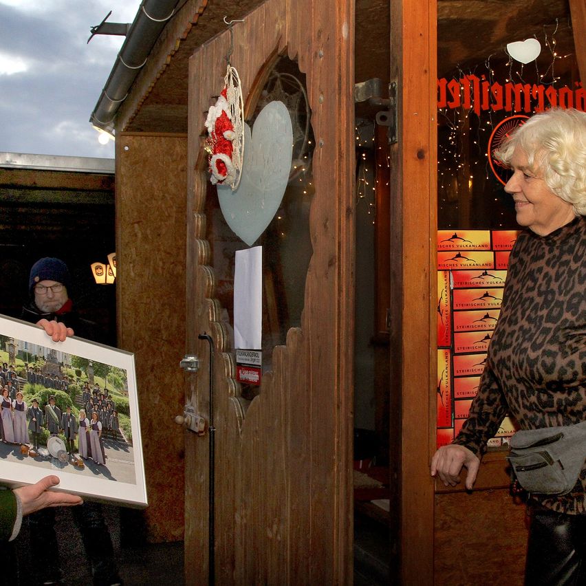 Eine Person hält ein Bild vor einer Holztür, eine Frau lächelt in der Nähe. Die Tür hat ein herzförmiges Fenster und eine Girlande. Die Tür führt in einen Raum mit einem roten und gelben Schild.