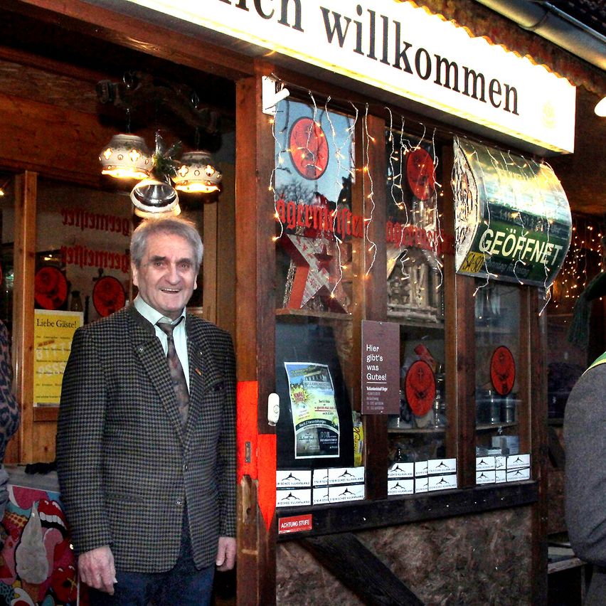 Ein Mann in einem Anzug steht vor einem Geschäft mit einem Schild, das 'Herzlich Willkommen' sagt. Das Geschäft ist mit Weihnachtslichtern geschmückt und hat Gegenstände im Schaufenster.