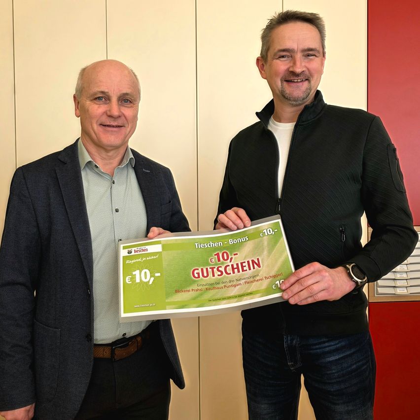 Zwei Männer stehen nah beieinander und halten einen grünen Gutschein, lächeln. Der Mann auf der linken Seite trägt einen grauen Anzug, während der Mann auf der rechten Seite einen schwarzen Mantel und Jeans trägt.