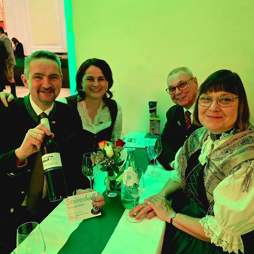 Vier Personen sitzen an einem Tisch und lächeln für ein Foto. Ein Mann hält eine Weinflasche und eine Frau ein Zertifikat. Hinter ihnen beleuchten grüne Lichter den Bereich.