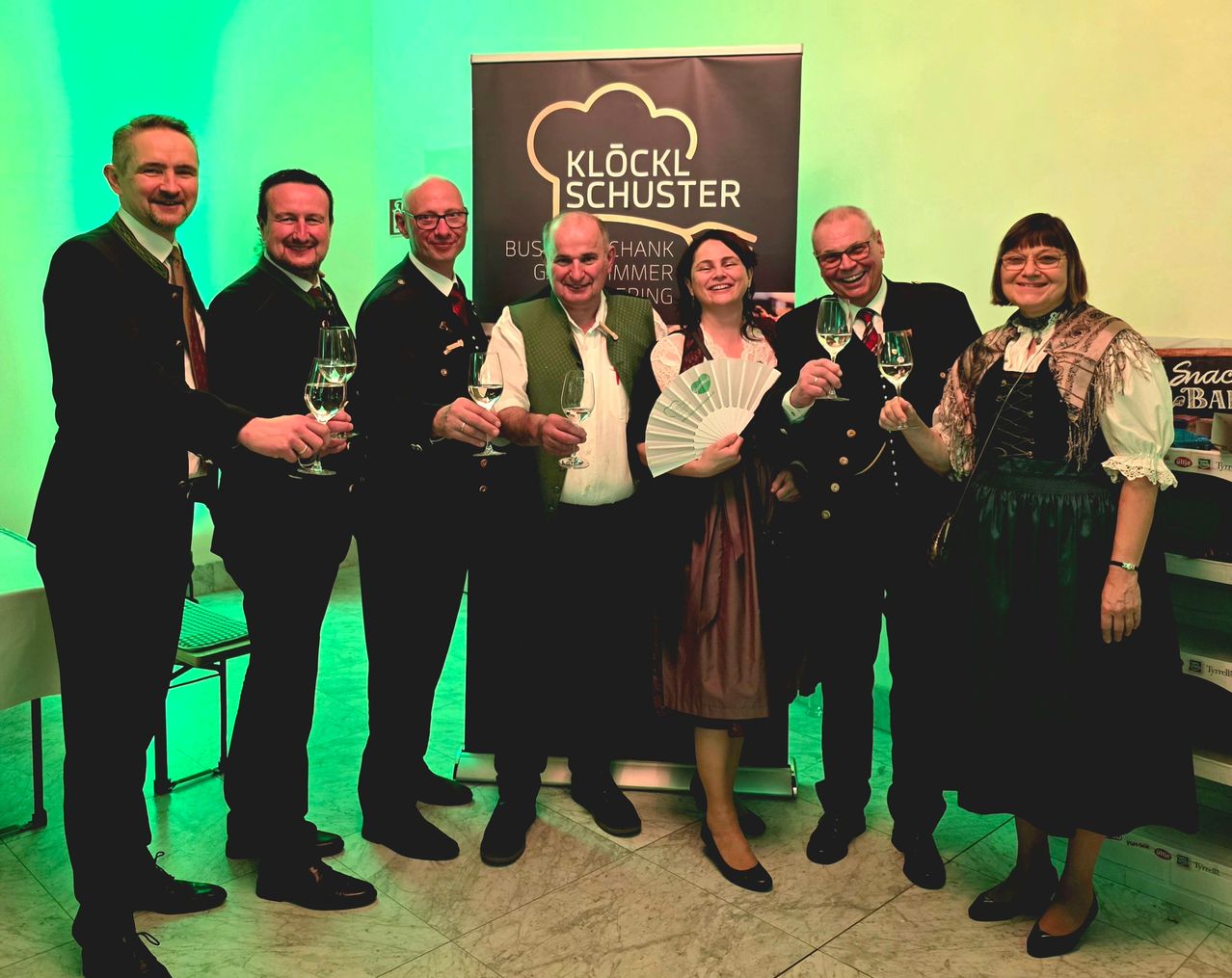 Eine Gruppe von Menschen in traditioneller Kleidung steht vor einem Banner. Sie halten Weingläser in der Hand und scheinen zu feiern. Sie lächeln und scheinen für ein Foto zu posieren. Der Hintergrund ist grün.