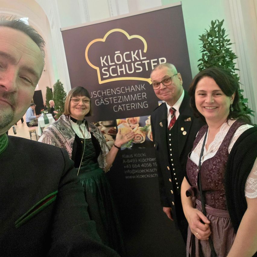 Vier Personen posieren vor einem Schild von Kloster Schuster. Zwei Frauen und ein Mann tragen traditionelle deutsche Kleidung. Hinter ihnen bewirbt ein Schild Catering-Dienstleistungen.