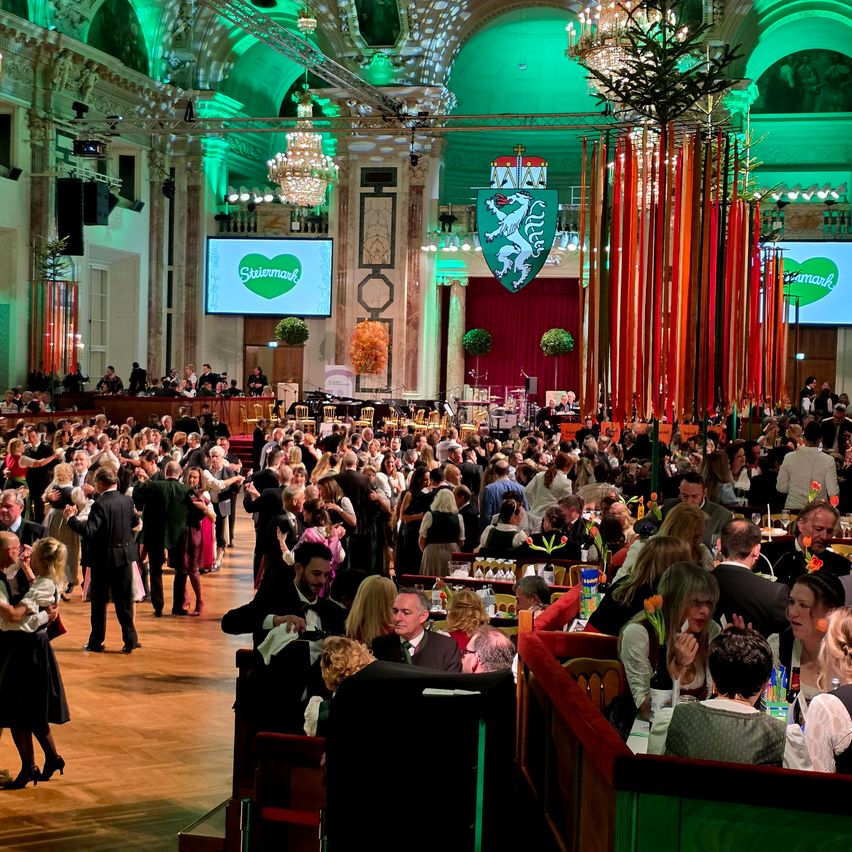 Eine große Indoor-Veranstaltung mit vielen tanzenden und an Tischen sitzenden Personen, geschmückt mit grünen Lichtern, Bannern und einer zentralen Bühne mit einer großen Leinwand, auf der das Wort 'Steiermark' steht.
