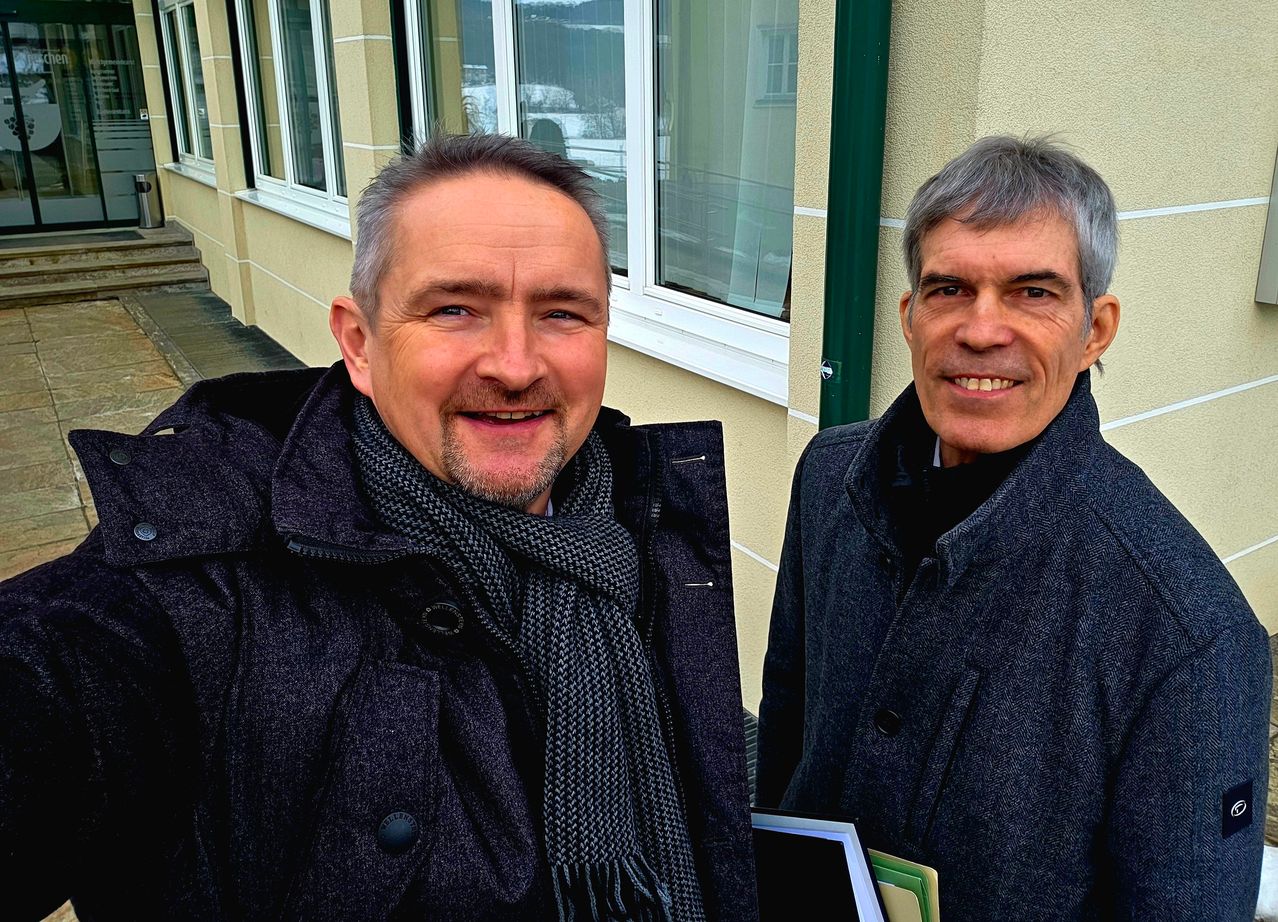 Zwei Männer stehen vor einem beigen Gebäude mit Glasfenstern. Einer trägt einen Schal, der andere hält eine Mappe. Sie lächeln beide.