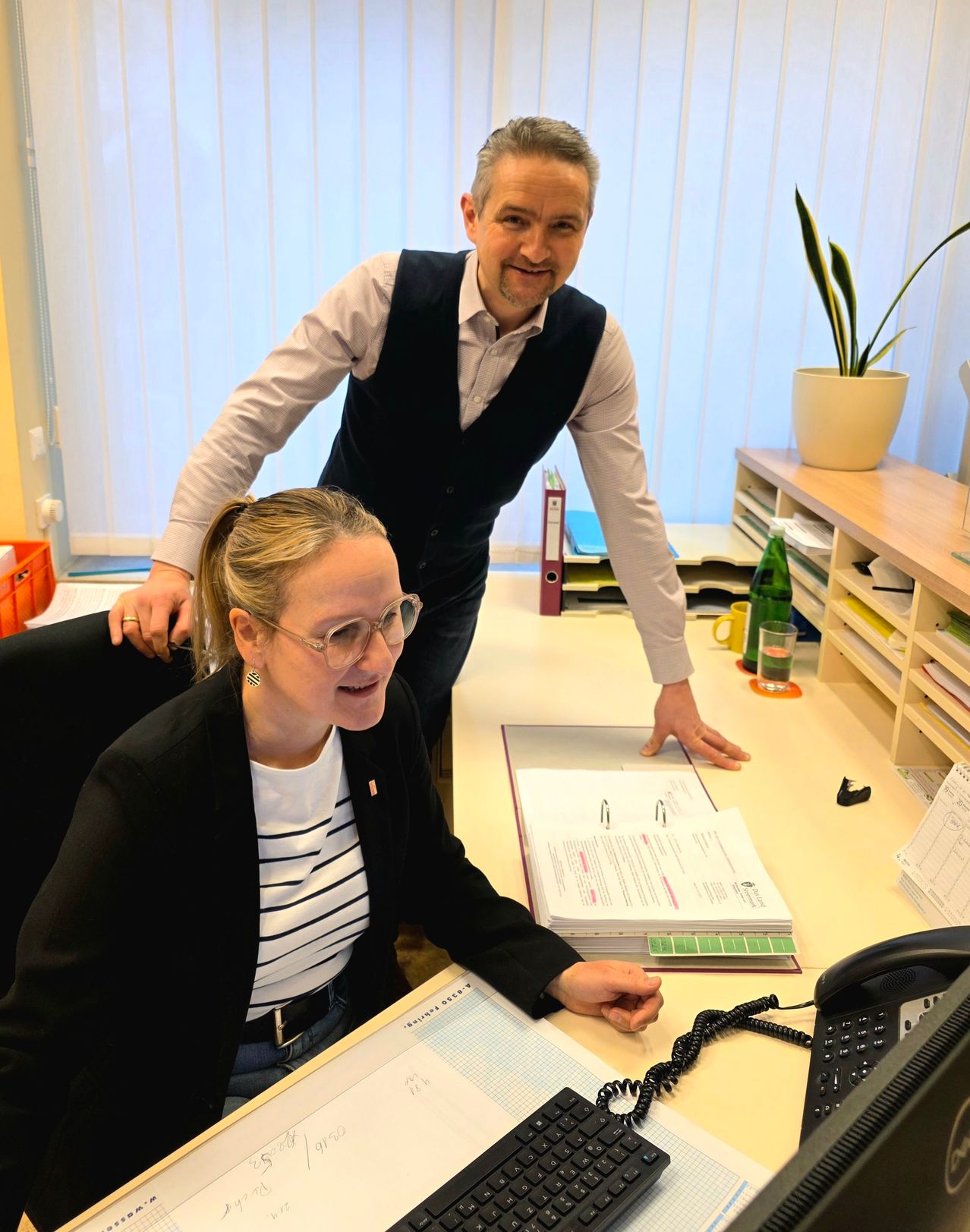 Ein Mann steht hinter einer Frau an einem Schreibtisch. Sie lächelt und er hat eine Hand auf ihrer Schulter. Der Schreibtisch ist mit Papieren, einem Telefon und einer Pflanze übersät.