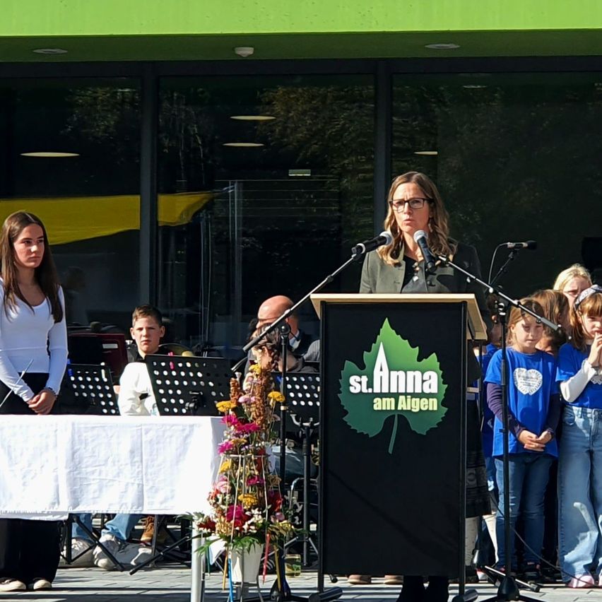 Eine Frau spricht an einem Podium mit Mikrofon. Hinter ihr steht eine Gruppe von Kindern aufmerksam. Vor dem Podium befindet sich ein Tisch mit Notenständern und einer Blumenarrangement.