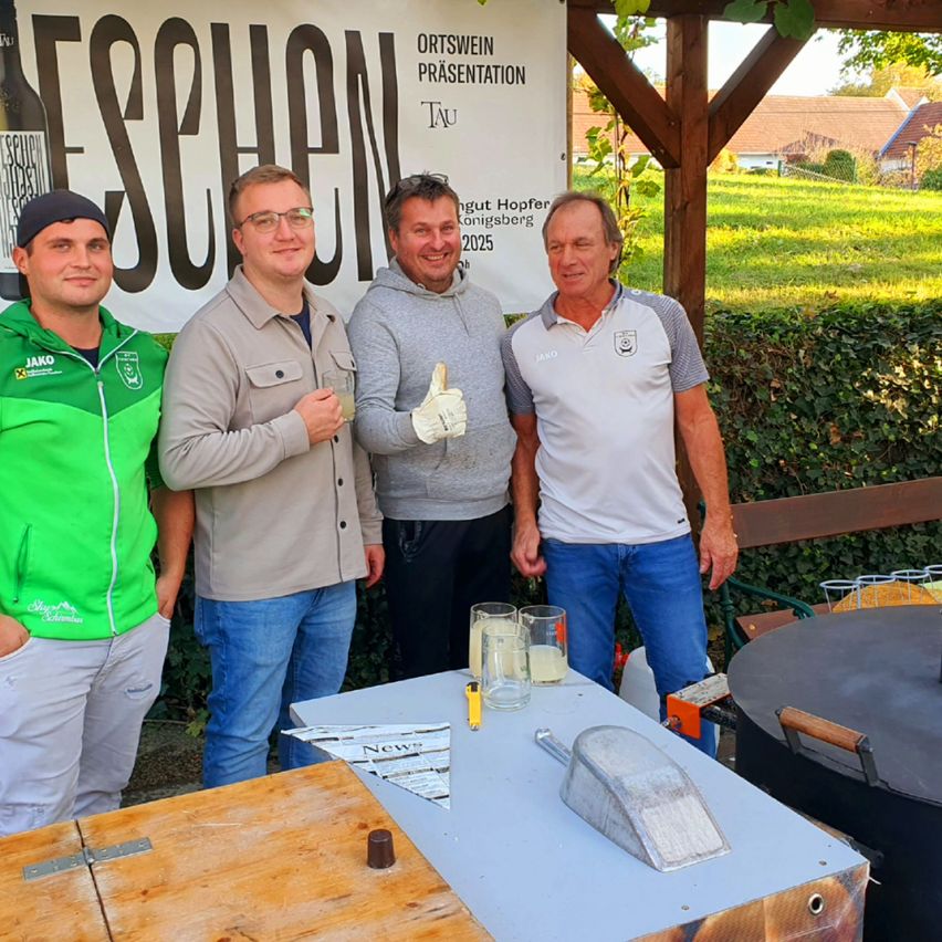 Vier Männer stehen bei einer Outdoor-Veranstaltung zusammen, halten Getränke in den Händen und lächeln. Hinter ihnen steht ein Schild mit dem Wort 'Taschen' und der Zahl '2025.' Sie befinden sich in der Nähe eines Tisches mit einem großen Topf und Gläsern.