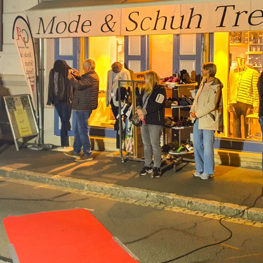 Einige Personen stehen vor einem Geschäft namens Mode & Schuh Tre. Eine Person fotografiert eine Jacke an einem Mannequin. Im Inneren sind Schuhe auf Regalen ausgestellt.