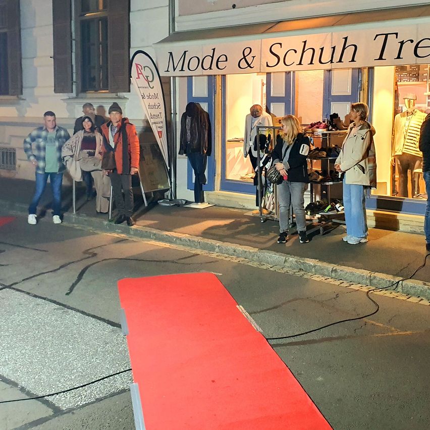 Eine Gruppe von Menschen steht vor einem Geschäft namens Mode & Schuh. Das Geschäft hat einen roten Teppich und ist nachts gut beleuchtet.