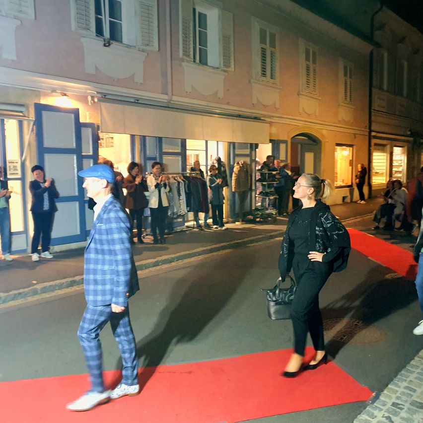 Ein Ereignis bei Nacht draußen, mit einem Mann und einer Frau, die auf einem roten Teppich gehen. Leute sind in der Nähe, einige bei Läden.