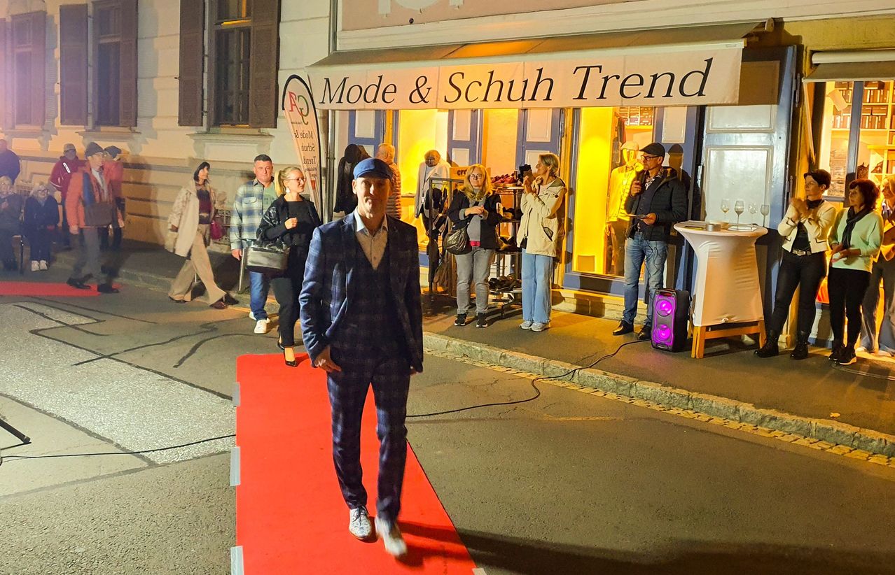 Ein Mann geht auf einem roten Teppich vor einem Geschäft namens Mode & Schuh Trend, während mehrere Leute im und vor dem Laden zusehen.