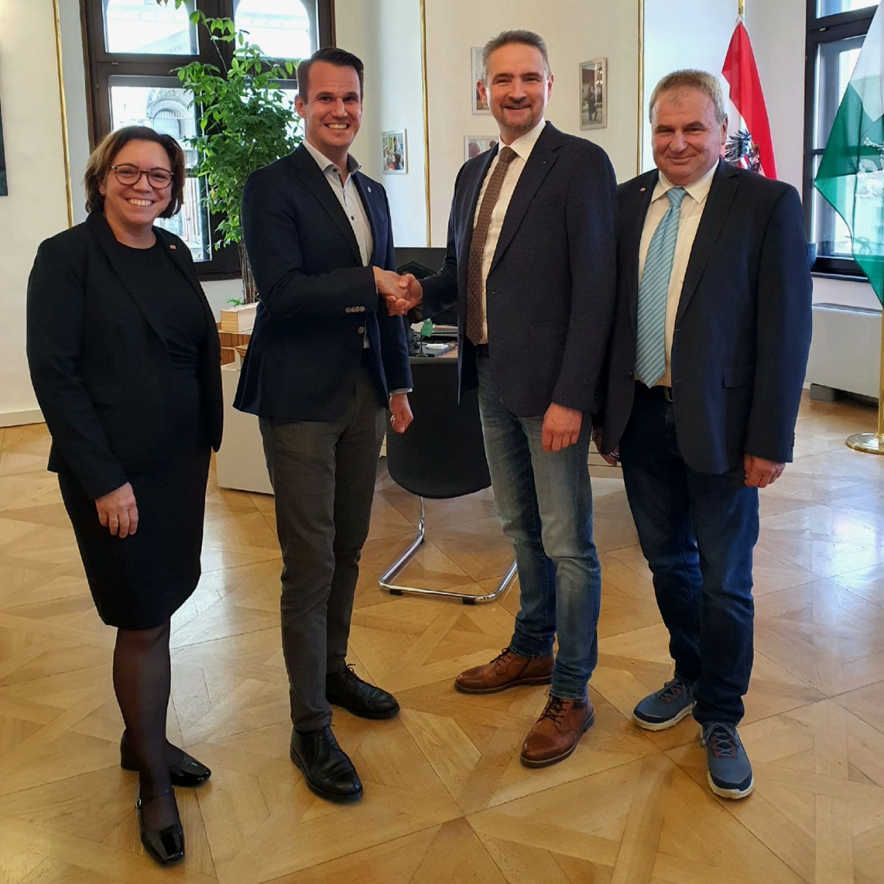 Vier Menschen stehen in einem Raum. Ein Mann schüttelt einem anderen die Hand. Zwei Flaggen sind im Hintergrund.