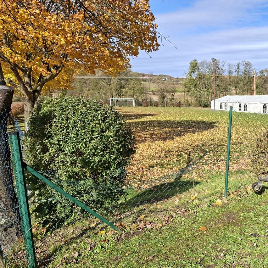 Eine Freiluftszene mit einem Grasfeld, verstreuten Blättern, einem Fußballtor und einem großen weißen Zelt mit Fenstern. Das Gebiet ist von Bäumen, Büschen und einem grünen Zaun umgeben.