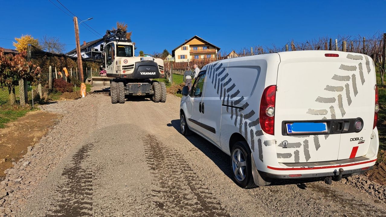 Ein weißer Lieferwagen mit Reifenspuren-Grafiken ist auf einer Schotterstraße in der Nähe eines Hauses mit Weinberg geparkt. Ein Mann steht neben einem Baggerlader.