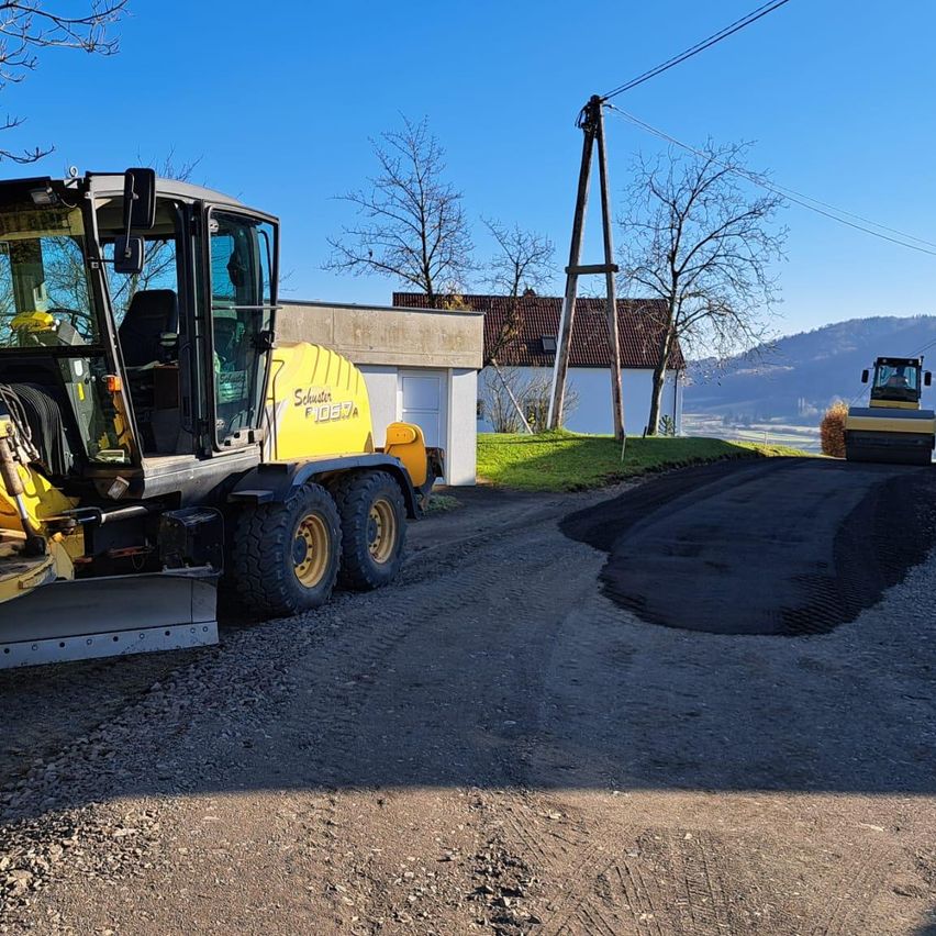 Zwei gelbe Baufahrzeuge stehen auf einer Schotterstraße. Ein Haus mit braunem Dach und ein Versorgungsmast stehen dahinter. Bäume und Berge im Hintergrund.