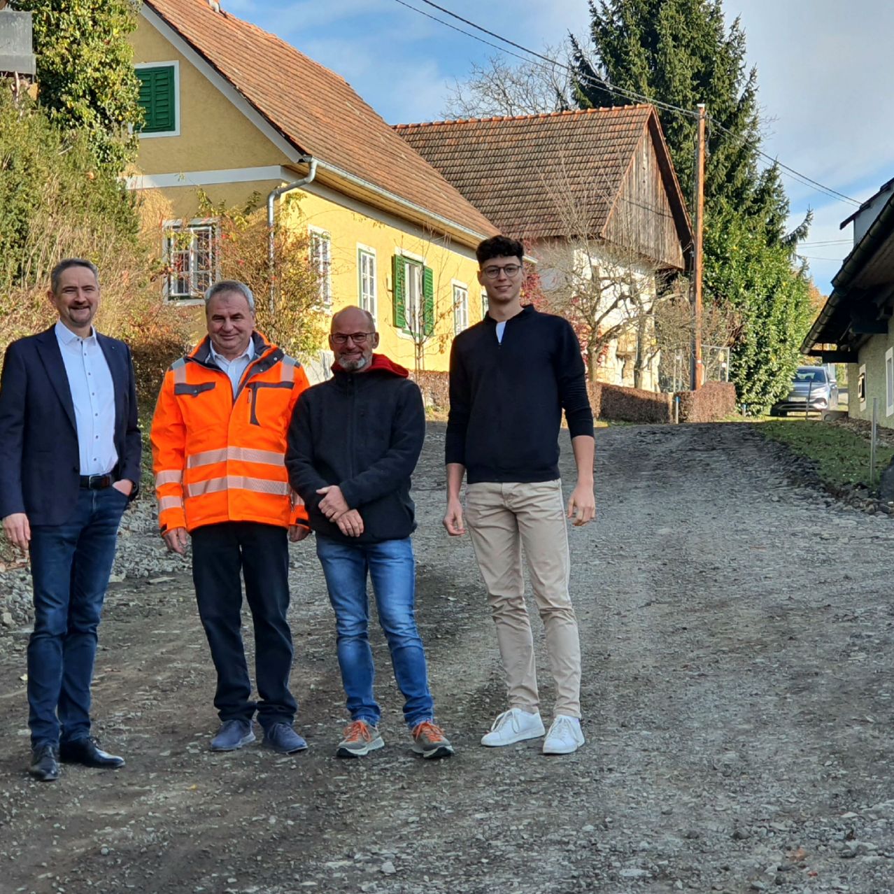 Vier Männer stehen auf einer Kiesstraße vor einem gelben Haus mit grünen Fensterläden und einem roten Dach. Die Männer sind in Geschäftskleidung und Freizeitkleidung gekleidet.