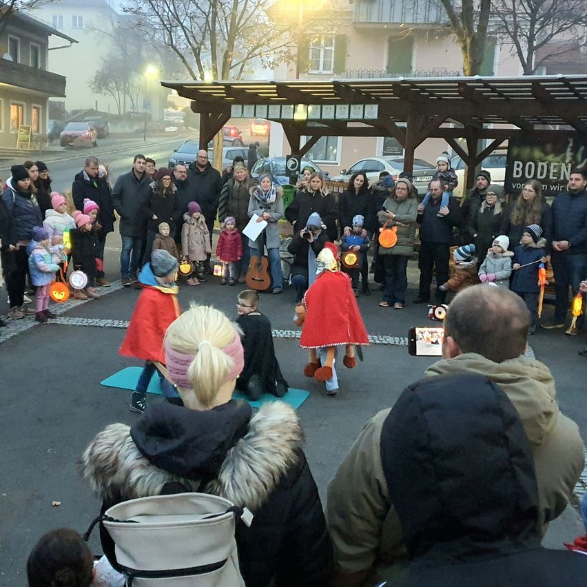 Eine Menschenmenge versammelt sich im Freien für eine Veranstaltung, einige halten Laternen. Zwei Kinder in roten Umhängen knien, während andere dahinter stehen. Ein Vordach und Bäume sind im Hintergrund.