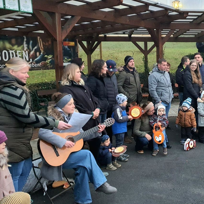Eine Gruppe von Menschen steht unter einem Pavillon. Eine Frau hält ein Papier und eine Gitarre, und ein Mädchen spielt die Gitarre. Einige Kinder und Erwachsene sitzen und hocken auf dem Boden.