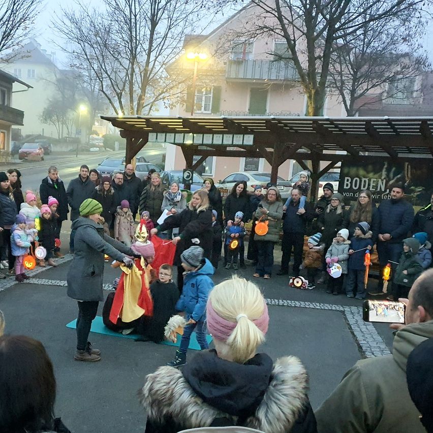 Eine Gruppe von Menschen, einschließlich Kindern, versammelt sich in einem nebligen Außenbereich. Einige sind in Kostümen gekleidet und halten brennende Laternen. Eine Frau kniet mit einem Kind, das eine Maske trägt.