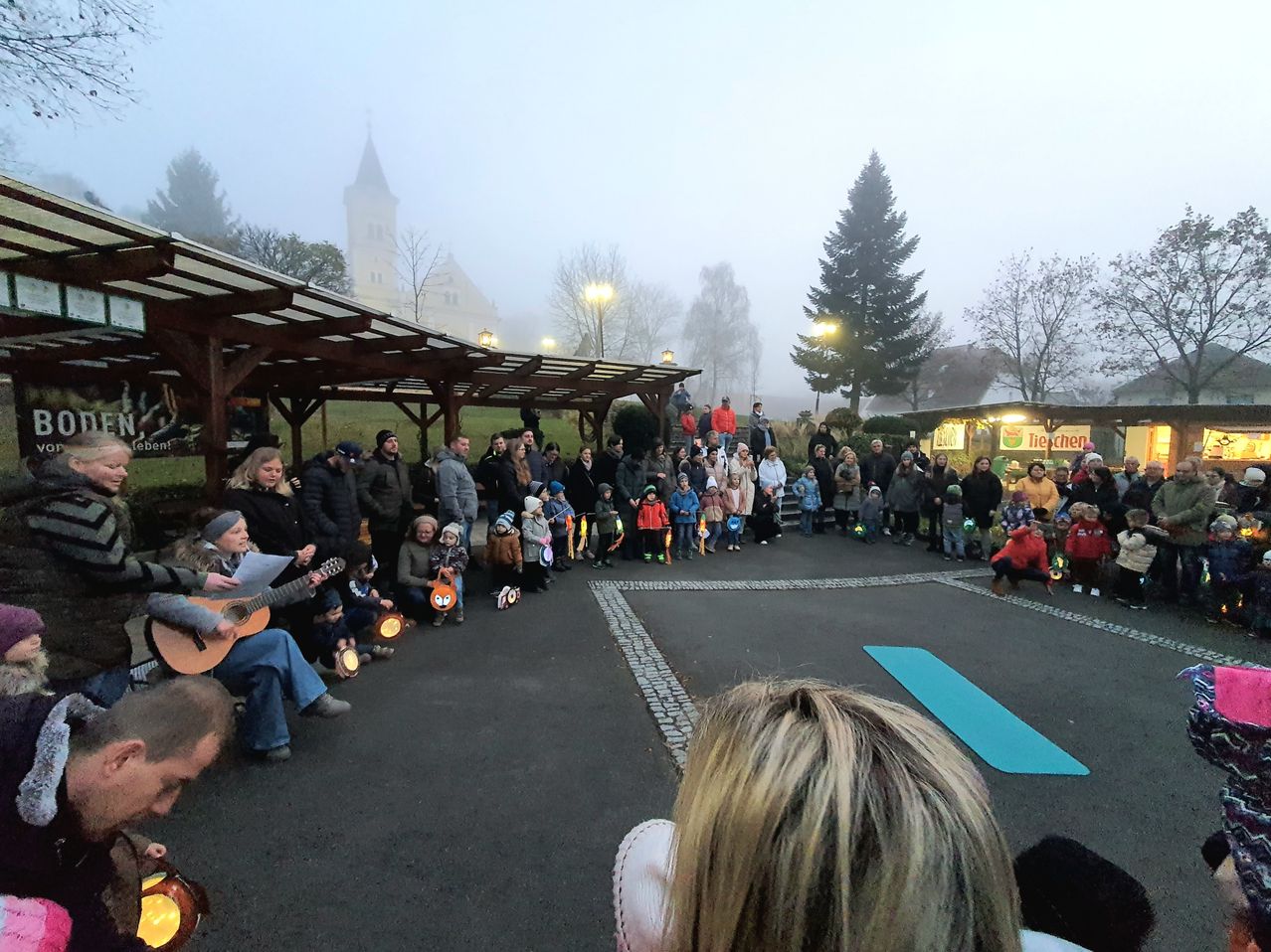 Eine Menschenmenge versammelt sich auf einem nebligen Außengelände, einige sitzen, andere stehen, und Kinder halten Laternen. Bäume, Straßenlaternen und ein Gebäude im Hintergrund.