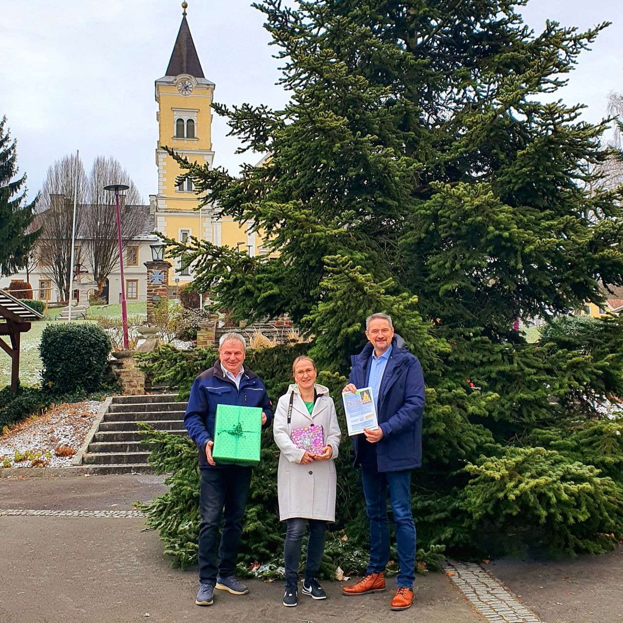 Drei Personen stehen vor einem hohen Tannenbaum und halten Geschenke. Hinter ihnen befindet sich ein Gebäude mit einem Uhrturm und einem Kirchturm.
