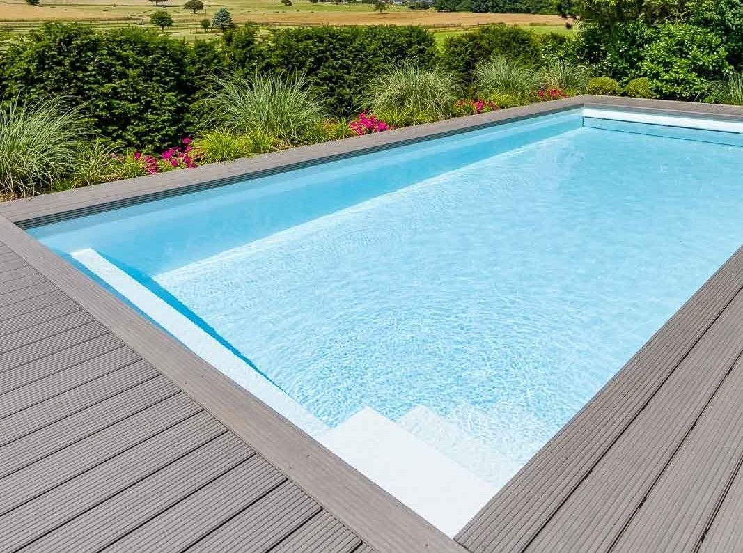 Ein kristallklarer, leerer Swimmingpool mit Holzterrasse, umgeben von einer üppigen grünen Landschaft mit blühenden Pflanzen.