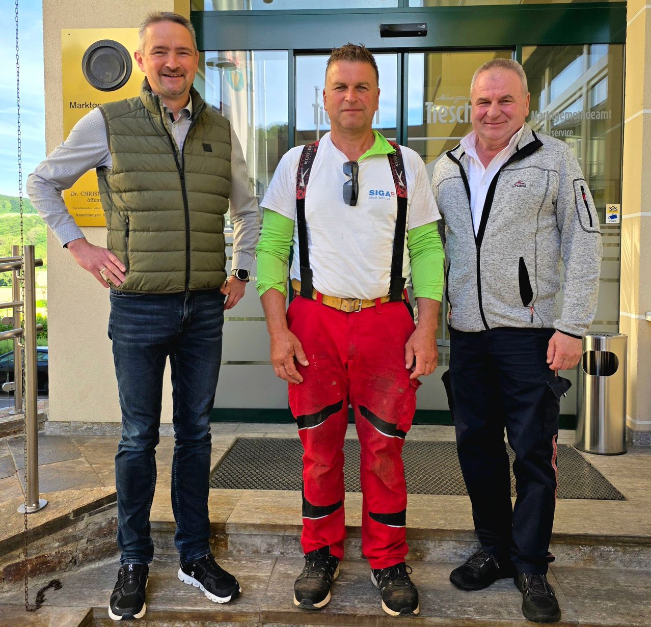 Drei Männer stehen lächelnd auf den Stufen vor einem Gebäude. Der Mann in der Mitte trägt ein weißes Hemd mit roten Hosen und einer Hosenträger. Der Mann links von ihm trägt eine Weste und Jeans. Der Mann rechts von ihm trägt eine graue Jacke. Hinter ihnen ist eine Glastür mit einem Schild.