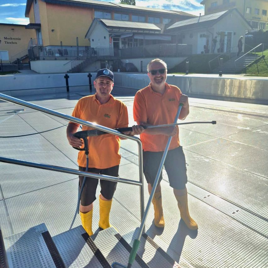 Zwei Männer in orangefarbenen Hemden und gelben Stiefeln halten Reinigungswerkzeuge auf einer Plattform mit einer Treppe. Gebäude mit Schildern sind im Hintergrund.