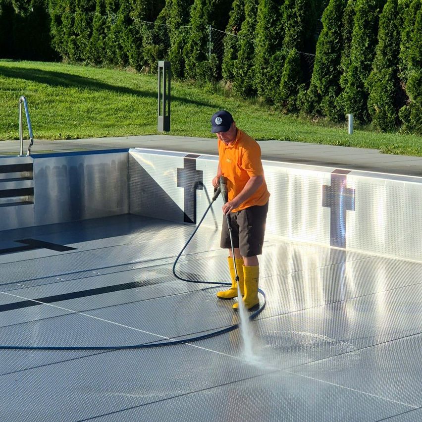 Ein Mann in einem orangefarbenen Shirt und gelben Stiefeln sprüht mit einem Schlauch Wasser auf eine Poolterrasse.