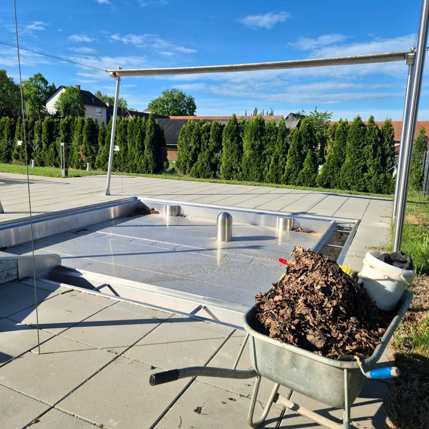 Eine Metallstruktur mit einem Metallgitter befindet sich auf einer Betonoberfläche. Ein Schubkarren mit getrockneten Blättern ist in der Nähe, und ein weißer Eimer mit einigen Blättern steht rechts. Dahinter befindet sich eine Rasenfläche mit Hecken und Gebäuden.