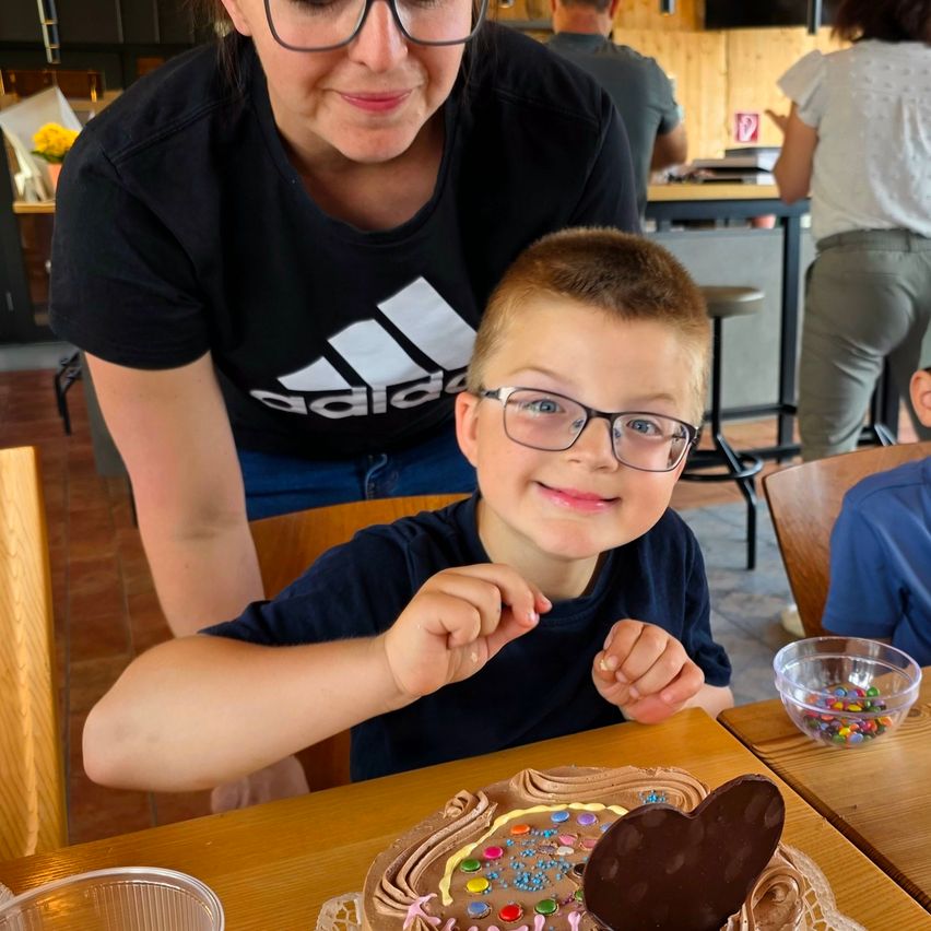 Ein junger Junge mit Brille lächelt in die Kamera, sitzt an einem Tisch mit einem Schokoladenkuchen. Eine Frau in einem Adidas-Shirt steht hinter ihm.