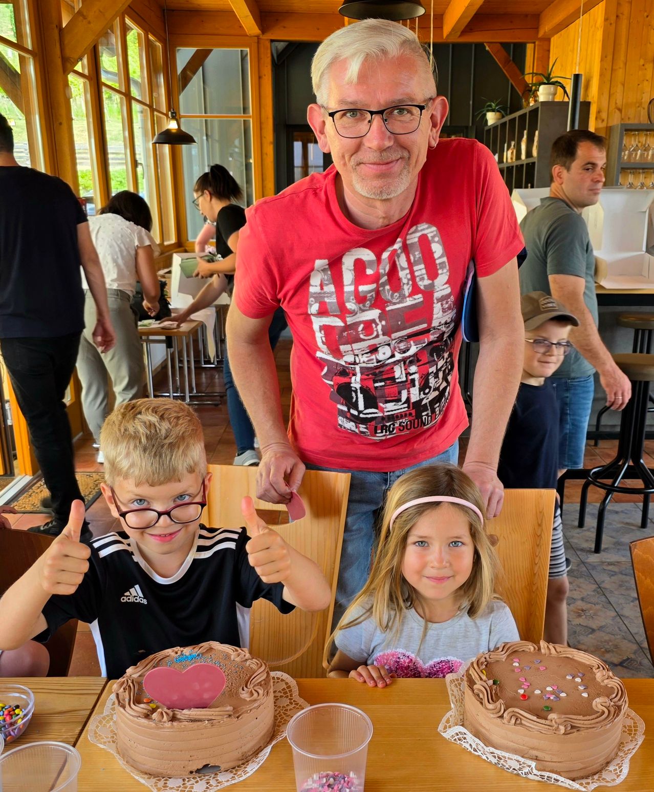 Ein Mann in einem roten T-Shirt steht hinter zwei Kindern in einem Restaurant. Beide Kinder lächeln und zeigen Daumen hoch. Auf dem Tisch stehen Schokoladenkuchen mit Herzformen.
