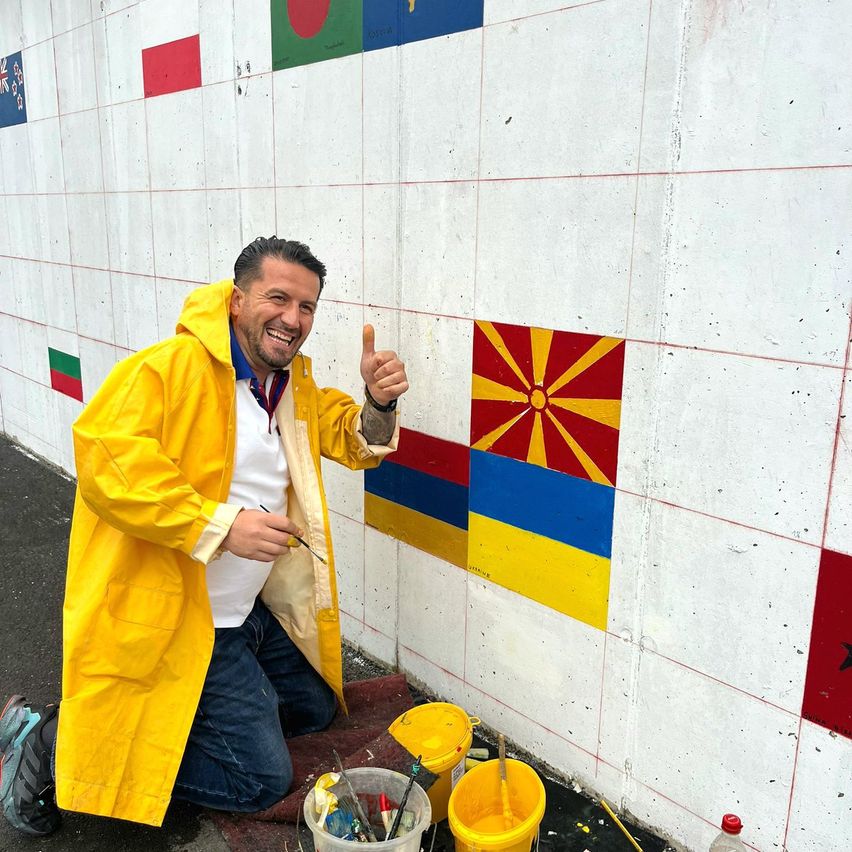 Ein Mann in einem gelben Regenmantel kniet am Boden und malt ein Wandbild mit Flaggen.