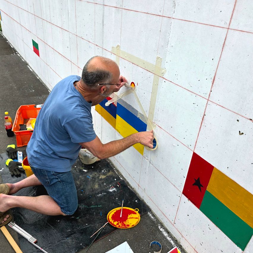 Ein Mann in einem blauen Shirt und Jeans kniet auf dem Boden und trägt Farbe auf eine Wand auf. Er ist von verschiedenen Werkzeugen und Farbdosen umgeben. Die Wand zeigt eine Reihe von Flaggen.