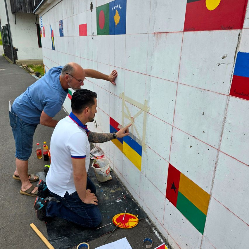 Zwei Männer malen Flaggen auf eine weiße Wand, einer zeigt auf die neue Malerei. Farbdosen, Pinsel und eine Palette sind in der Nähe.